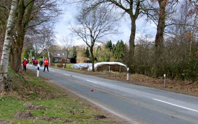 Boßelspieler wirft Kugel auf Landstraße, Gruppe beobachtet am Rand.